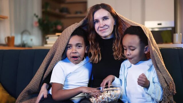 Camera view of young woman mother with little african american boys brothers eating popcorn on sofa under the blanket watching movie film show television at late night at home together Family time
