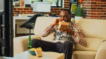 Modern guy eating cheeseburger with fries from takeout, enjoying fast food delivery meal and watching comedy movie on television. Young happy adult feeling relaxed in front of tv.