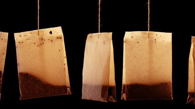 Set of used brown paper tea bags with tasty herbal brew hang swaying on thin threads on black background extreme close view