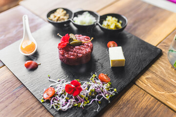 Elaborate smoked beef tartare dish served on black wooden board with garnishes and egg yolk. Polish cuisine. Restaurant table. Horizontal indoor shot. High quality photo