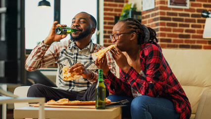 Obraz premium Young couple laughing and eating pizza, sitting on living room couch and enjoying fast food takeaway meal. Happy partners watching comedy movie on tv channel program. Handheld shot.