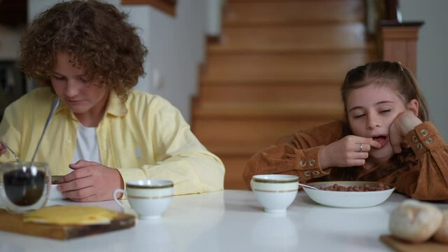 Front view portrait of schoolboy and schoolgirl eating morning cereal sitting at table in living room. Sleepy Caucasian girl putting head on table closing eyes as boy enjoying tasty breakfast