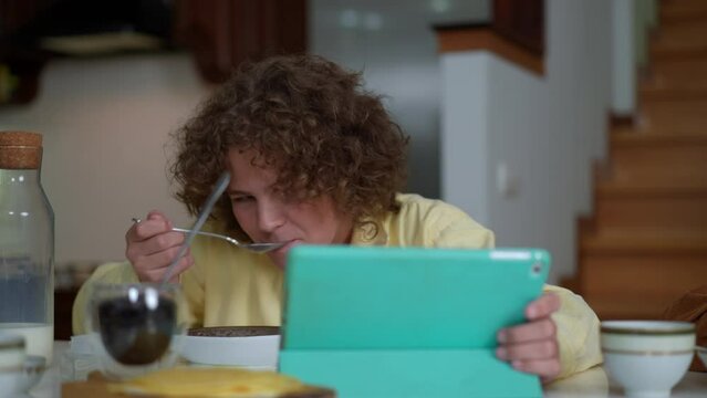 Concentrated boy eating morning cereal in slow motion watching online movie on digital tablet. Portrait of curios focused Caucasian schoolboy at home in the morning before school