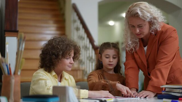 Serious Caucasian woman talking with children painting picture sitting at table indoors. Portrait of intelligent Caucasian mother helping son and daughter doing homework