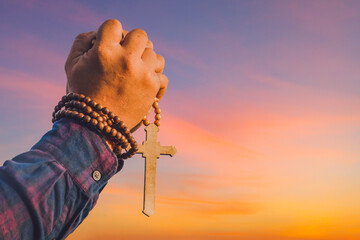Male hand holding wooden cross necklace with sunset sky background