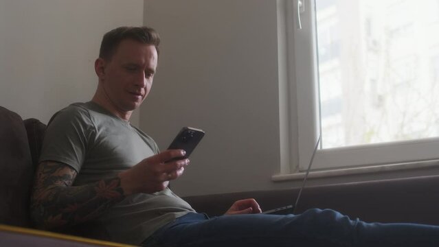 home office worker sit on couch with laptop on his lap and take call on phone. customer or boss call his executor, freelance programmer, web designer. Tattooed CEO talking on smartphone at home.