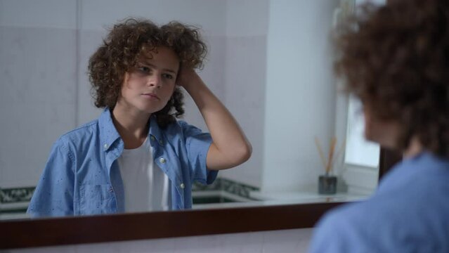 Reflection In Bathroom Mirror Of Concentrated Boy Touching Curly Hair Thinking. Cute Caucasian Schoolboy Adjusting Haircut Getting Ready For School In The Morning At Home Indoors