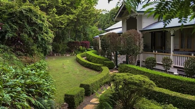 Smooth cinematic descending reveal shot,through lush foliage, of a classic traditional country Queenslander house, nestling in neatly manicured landscaped garden