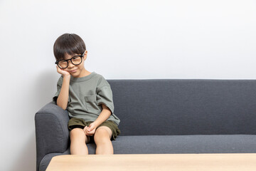 Cute Asian boy with eyeglasses looking up or right, sitting on a sofa at home. Curious kid thinking.