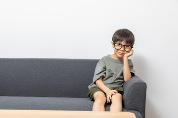 Cute Asian boy with eyeglasses looking up or right, sitting on a sofa at home. Curious kid thinking.