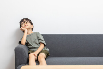 Cute Asian boy with eyeglasses looking up or right, sitting on a sofa at home. Curious kid thinking.