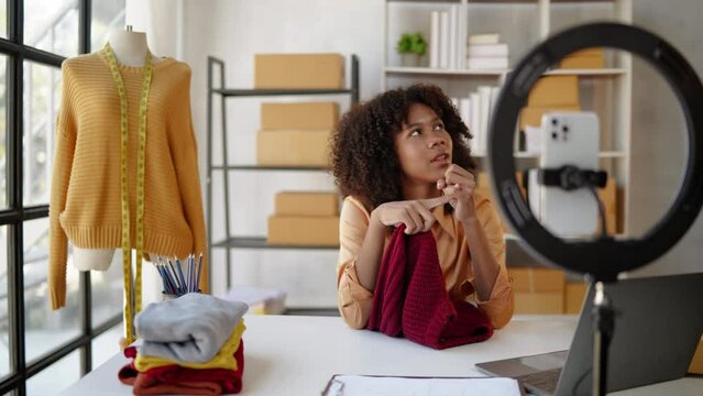 Female African designer and online seller is live streaming broadcast for selling new clothes and receiving the order from customer  and packaging product in workspace.