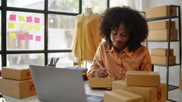 Female African designer and online seller is receiving new clothes orders from customers on laptop and packaging products into parcel boxes in workspace.