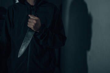 A male murderer with a knife in a dark room looks terrifying as he is about to injure the horror victim of a bloodthirsty killer holding a huge bloody kitchen knife in his hand.