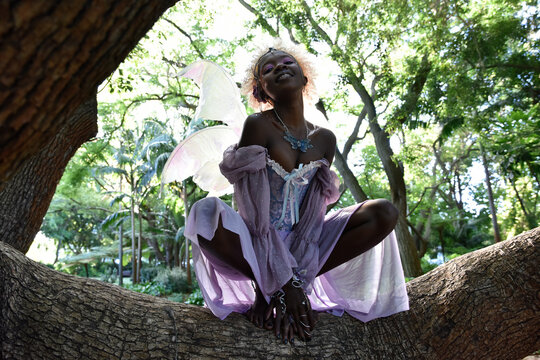 Portrait of beautiful African woman wearing purple fantasy costume, magical fairy wings & flower crown afro, climbing tree branches in forest location with natural glowing lighting. - Powered by Adobe
