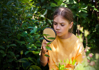 girl sits in the bushes outdoors in summer and looks through a magnifying glass
