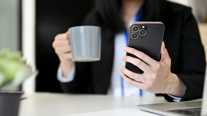 Fototapeta premium Close-up image of a businesswoman using her smartphone and sipping coffee at her office desk.