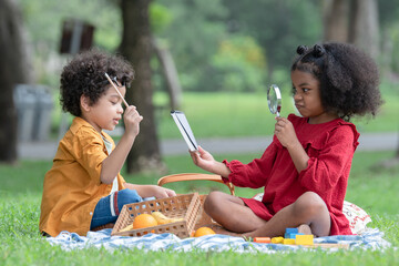Little cute African children boy and girl sitting and have fun playing magnifying glass to read book while picnic at park together. Sibling relationship in family. Lifestyle and education concept