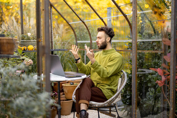 Man works on laptop, talking online while sitting by the round table in glasshouse with plants and flowers at backyard. Work from home at cozy atmosphere on nature