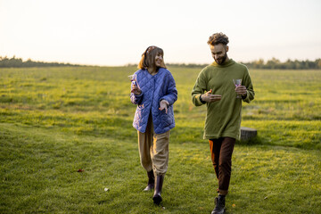 Stylish man and woman dressed warmly have conversation while walk together with wine on green lawn during a sunset. Couple spend leisure autumn time on nature