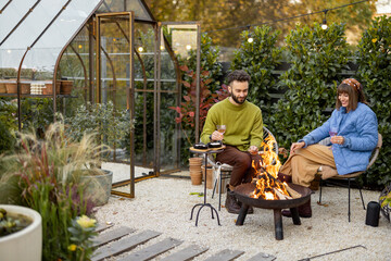 Young stylish couple grilling food and warming up while sitting together by the fire, spending...
