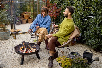 Young stylish couple grilling food and warming up while sitting together by the fire, spending...