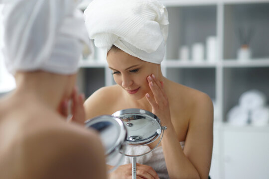 Young woman sits in the bathroom in front of the makeup mirror and does cosmetic procedures. Beautiful girl in white towel. Skin care, health, rejuvenation and spa treatment concept.