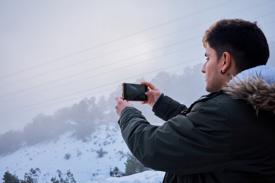 A young latin man takes a photo with his phone surrounded by snow