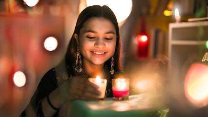 Obraz na płótnie Canvas Smiling pretty Hindu girl wearing big earrings sitting look hold burning glass candles closer to face, Indian teen enjoy celebrate Deepavali festival by lighting Diyas with colorful background