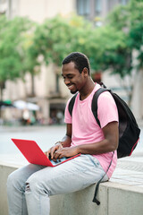 Man with backpack smiling while using a laptop sitting on a bench outdoors.