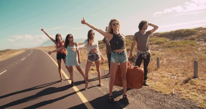 Hipster multi-ethnic group of friends hitchhiking on two lane highway in the sunshine with thumb sticking out and suitcase in hand