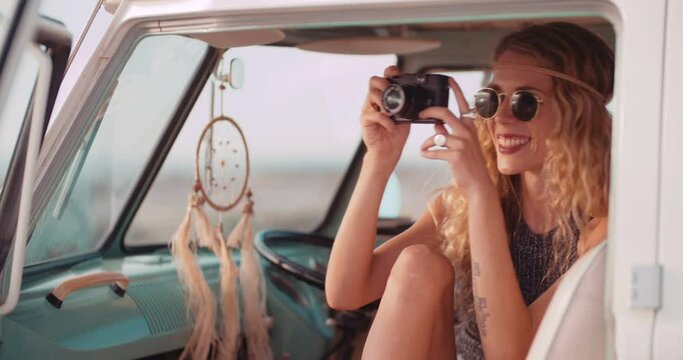 Road Trip Blonde Boho girl taking a photo of the sunset from front seat of retro van parked on a sandy beach