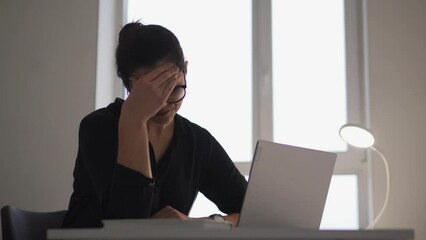 Concerned young woman working on laptop in office and feeling unpleasant pain in forehead, suffering migraine and high blood pressure, overworking