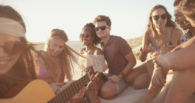 Group of friends relaxing at a sunset beachparty with someone playing a guitar in Slow Motion