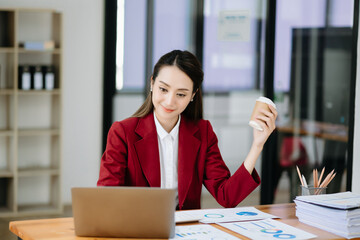 Confident beautiful Asian woman typing laptop computer and digital tablet while holding coffee at office.