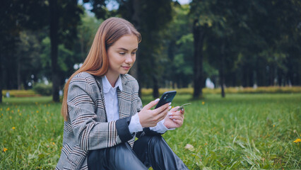A young girl makes a purchase with her phone and bank card in the park, sitting on the grass.
