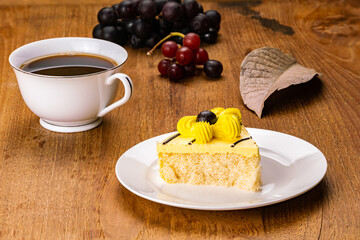 Breakfast with homemade delicious sponge cake garnish with yellow flower cream in white ceramic dish with a cup of black coffee and bunches of ripe grapes on wooden table.
