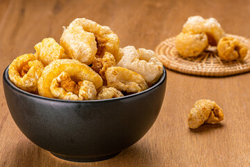 Closeup view of homemade crispy deep fried pork rinds in black ceramic bowl and on bamboo mat on wooden background.