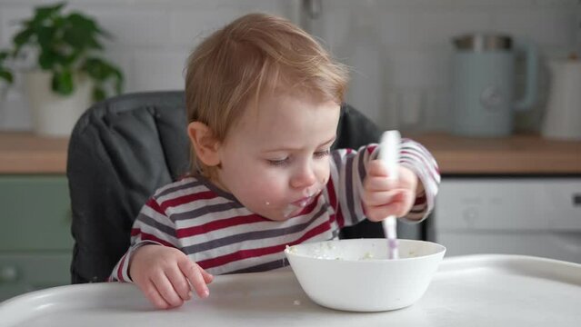 One year old hungry baby girl in striped casual clothes sits at white table in highchair and eats porridge herself with spoon. Blurred dining room background. Healthy eating for kids. Child nutrition