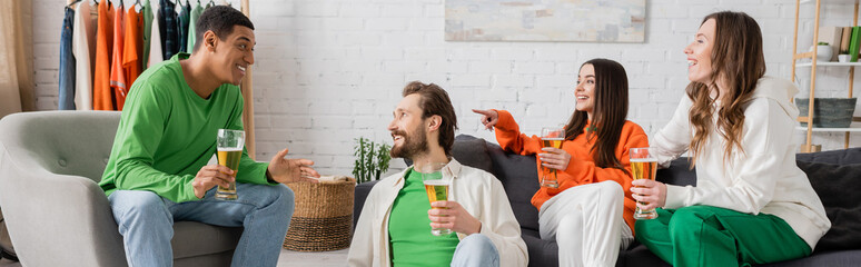 cheerful woman sitting near friend and pointing at interracial men talking in living room, banner