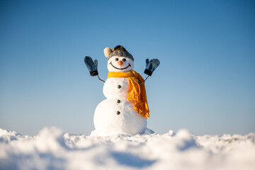 Funny snowman in knitted hat and yellow scalf with hands up on snowy field. Blue sky on background