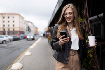 stylish young woman with a phone in her hands and a glass of coffee is walking down the street