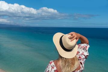Mujer rubia de espaldas, con un vestido floreado, sosteniendo un sombrero de paja en una mano, mirando el mar turquesa y cristalino de la costa de la tur&iacute;stica Fuerteventura en las islas Canarias