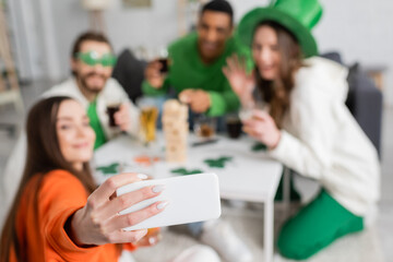 Blurred interracial friends taking selfie on smartphone while celebrating saint patrick day at home