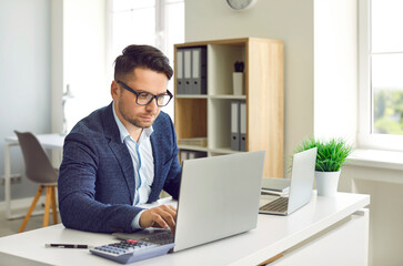 Concentrated male executive manager doing financial market research while working on laptop in office. Serious man in suit and glasses is sitting at table and working with laptop and calculator.
