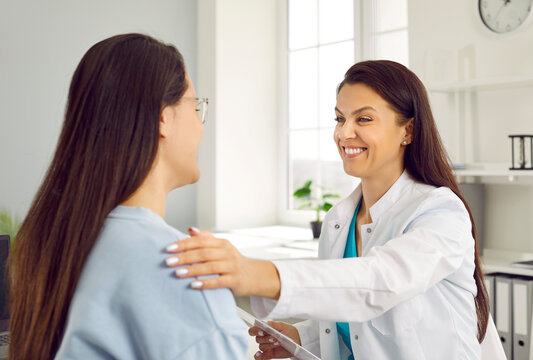 Friendly smiling female doctor holding woman's shoulder telling her positive news about her health. Woman in white coat welcomes female patient in her office. Concept of medicine.