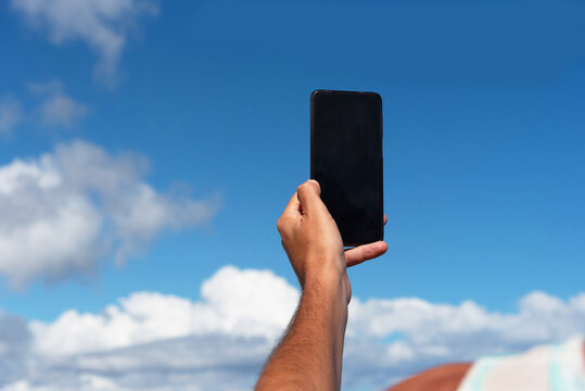 Primer plano del brazo de un hombre bronceado tomando una foto con su tel&eacute;fono m&oacute;vil del cielo azul con algunas nubes, durante un d&iacute;a soleado en las tur&iacute;sticas Islas Canarias.