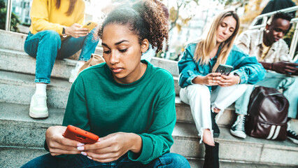 Multiracial young people using smart mobile phone device outside - University students looking smartphone sitting in college campus - Teenagers addicted to trendy technology and social media - obrazy, fototapety, plakaty