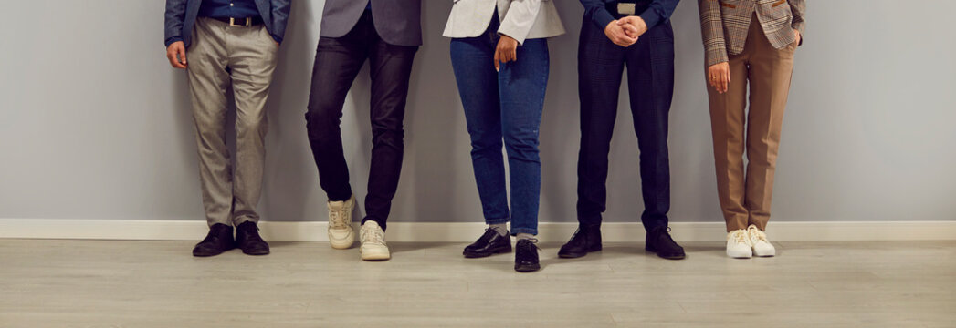 Business team standing in office. Diverse group of unrecognizable men and women in smart casual clothes of restrained colors standing by office wall. Cropped shot, people's legs. Banner background