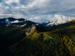 Drone landscape photo of the mountains in early Autumn. Some little clouds are flying low between mountains over it massive gray clouds and blue sky high. Sunlight breaking through the clouds.